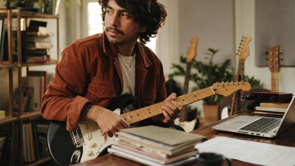 An independent artist working at a recording setup with a guitar and laptop, building out a body of work over time