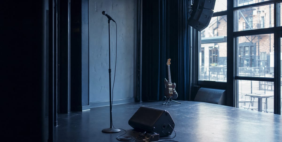 An empty small venue stage with a microphone, wedge monitor, cable, and a guitar resting on a stand in cool blue-grey light, no readable text.