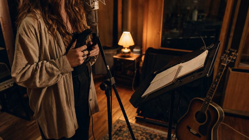Vocalist at a vintage microphone in a warm-lit studio with handwritten lyric drafts on a music stand.