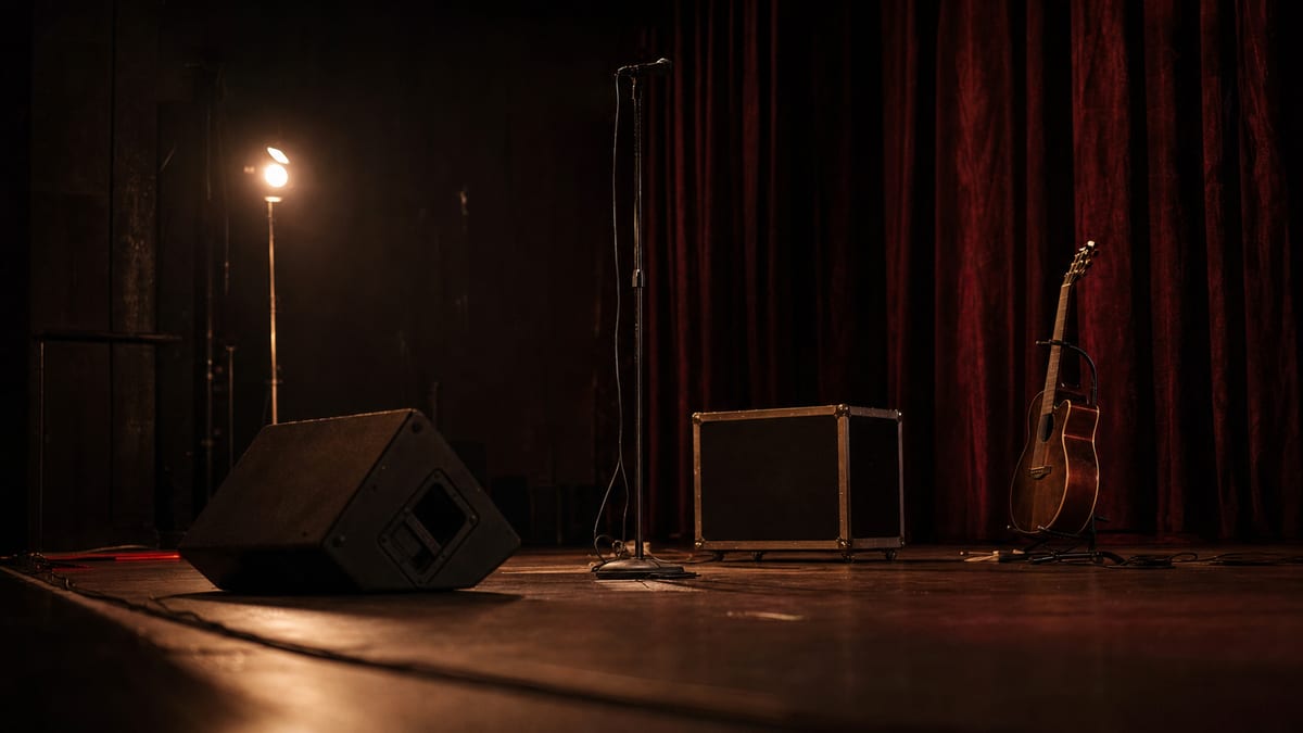 Dark rust-toned small venue stage before soundcheck with a microphone stand, monitor wedge, closed road case, cables, and a resting guitar.