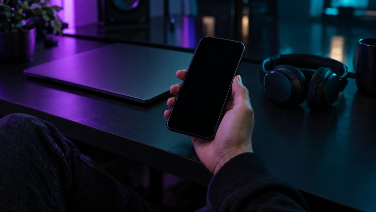 A minimal blue-gray studio desk with a phone showing a completely blank dark screen, a pair of headphones, and a closed notebook with no visible markings.