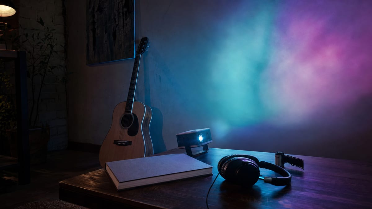 Dark studio corner with a resting acoustic guitar, closed sketchbook, small projector, headphones, and teal violet light.