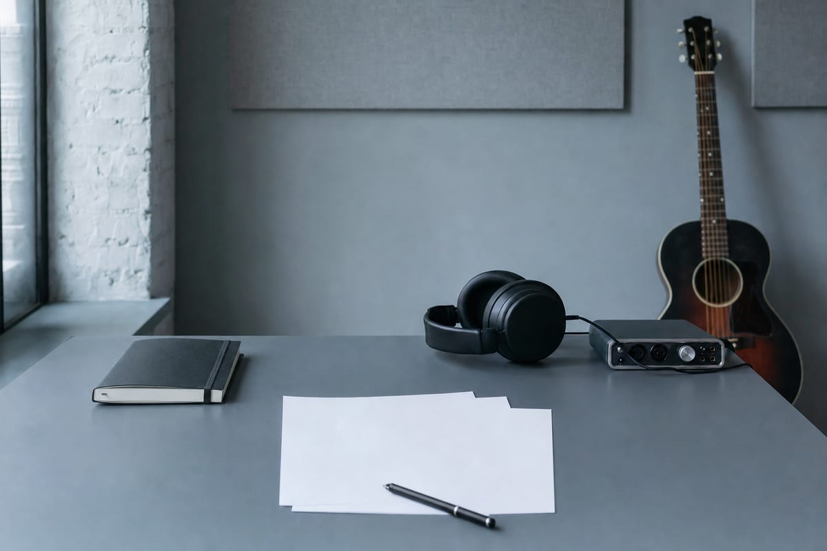 Cool blue-gray studio desk with a closed notebook, blank papers, headphones, audio interface, and a guitar resting against the wall.
