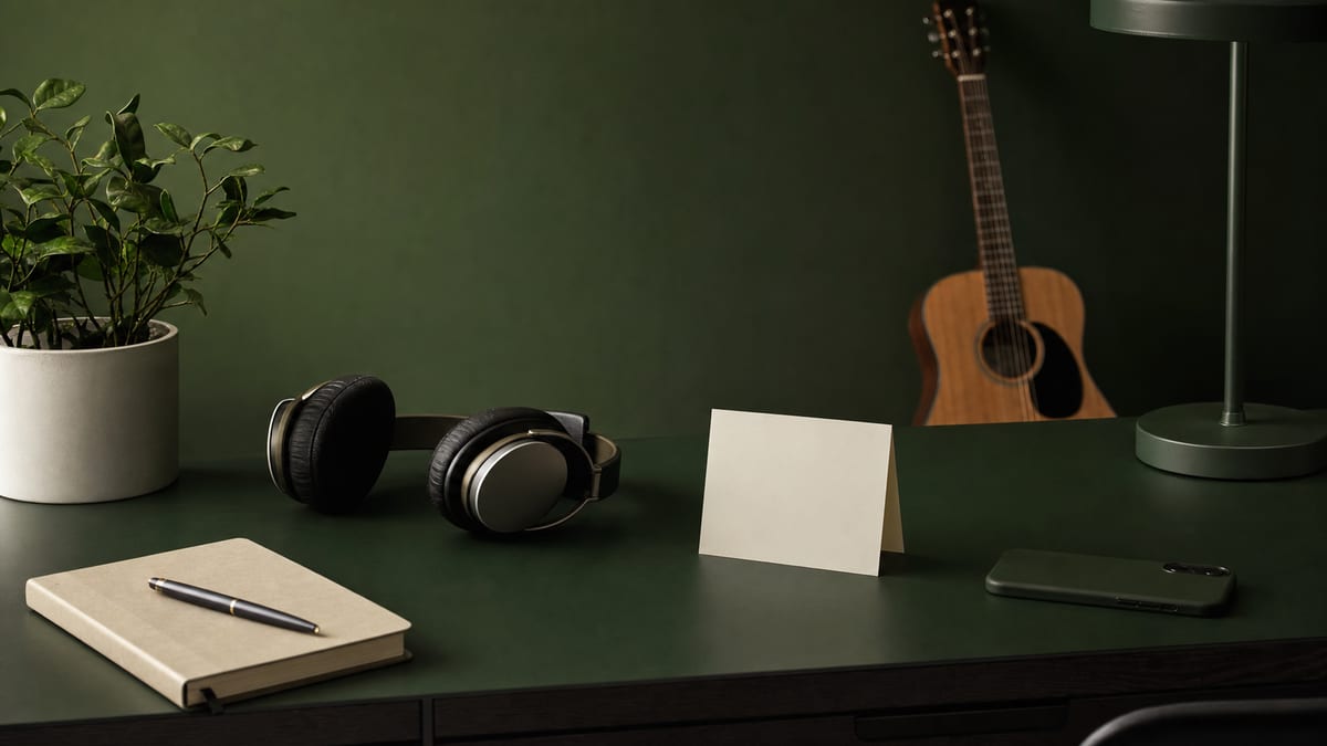 Minimal dark green and cream desk with headphones, blank release card, phone turned face down, plant, and guitar resting in the background with no readable text or logos.