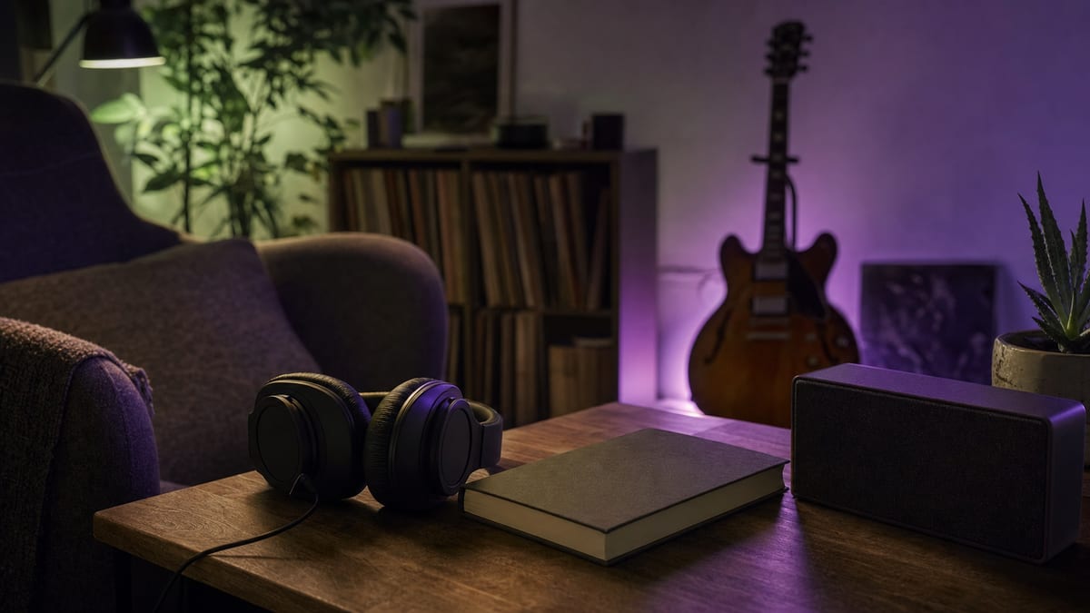 Warm listening corner with unbranded headphones, speaker, blank notebook, and a guitar resting on a stand under green and violet light, with no readable text or logos.